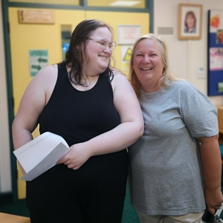 Two ladies one being a adult learner standing together indoors, with one holding a white paper with her gcse results on