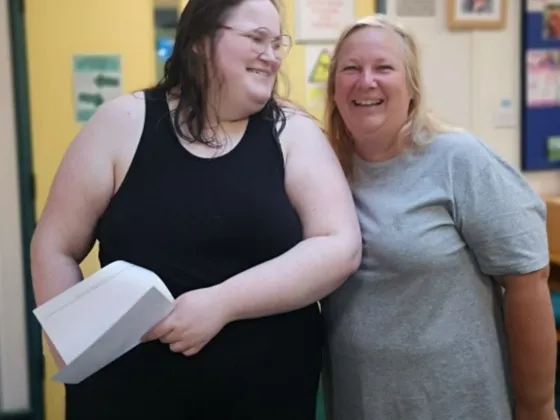 Two ladies one being a adult learner standing together indoors, with one holding a white paper with her gcse results on
