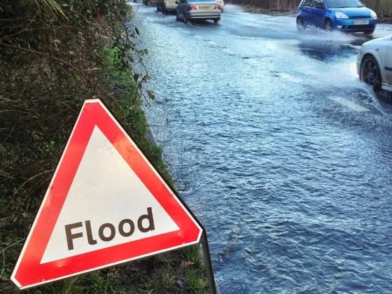 Flood warning sign beside a partially flooded road with cars driving through water and trees lining the roadside