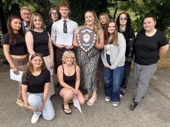 A group of people pose outdoors. Two people in the centre are holding an award shield and a certificate, while others stand or kneel around them.