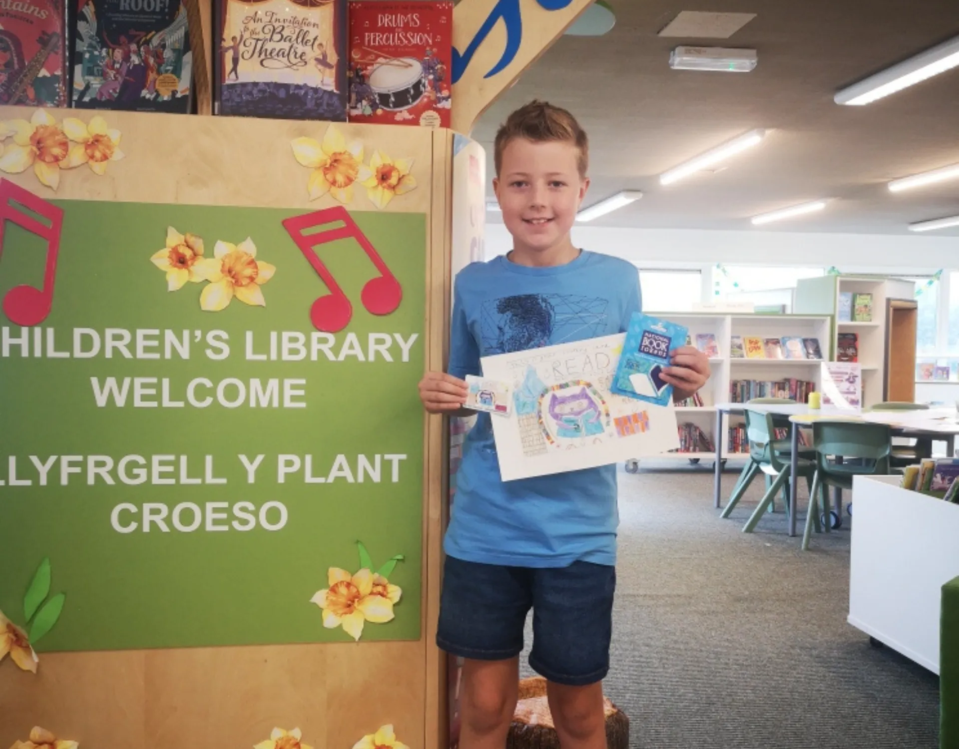 A young boy holding up his library card competition design and his new library card at Cwmbran Library.