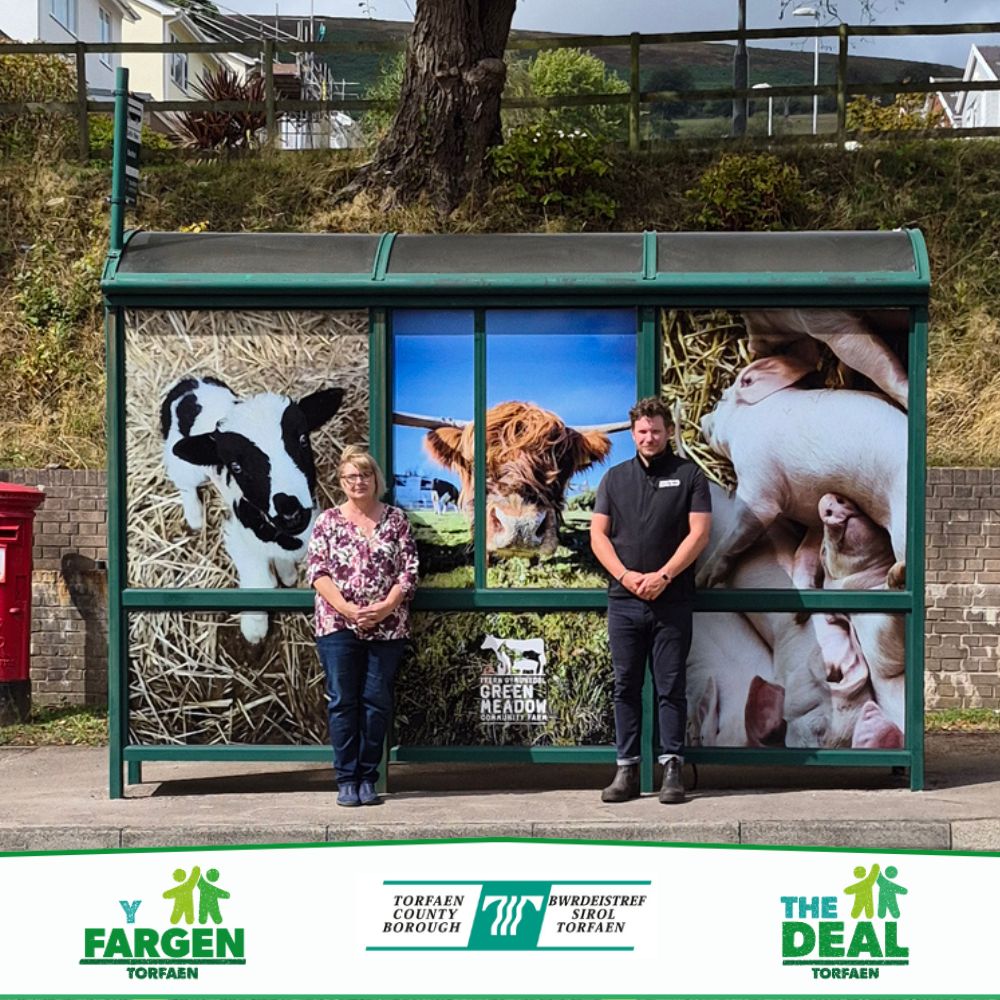 Cllr Owen and Greenmeadow Community Farm manager Jac Griffiths standing outside a bus stop which has been wrapped in farm animal imagery